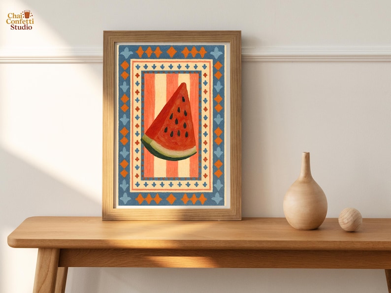 May include: A framed print featuring a watercolor illustration of a watermelon slice. The artwork has a red, orange, and blue border with a striped background. The print is displayed on a wooden table.