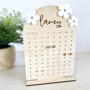 May include: A wooden earring holder with the name "Laney" in script. The holder has multiple holes for earrings and is decorated with white flower cutouts. Several pairs of earrings are displayed.