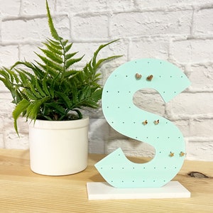 May include: A light blue wooden earring holder shaped like the letter "S" with a white base. The holder has multiple holes for earrings and is decorated with small white dots. There are several gold earrings on the holder.
