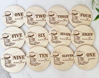 May include: Twelve wooden baby milestone discs with a cowboy boot design. Each disc features a different number from one to twelve, with the words "months" or "year" printed below.