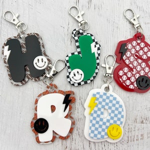 May include: Five colorful letter charms with a smiley face and lightning bolt design. Each charm has a different pattern, including basketballs, baseballs, checkered, and stripes. The charms are attached to silver key rings.