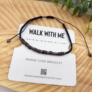 May include: A black Morse code bracelet with dark brown beads is displayed on a wooden surface. The bracelet is on a white card that reads "WALK WITH ME" and "MORSE CODE BRACELET". The card also includes a QR code and a website address.