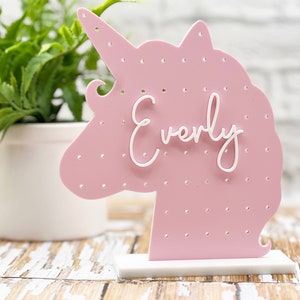 May include: Pink unicorn-shaped earring holder with white polka dots and the name "Everly" written in white. The holder has a white base.