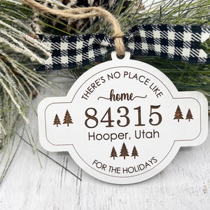 Puede incluir: Adorno navideño de madera blanca con el texto "THERE'S NO PLACE LIKE HOME" y "84315 Hooper, Utah FOR THE HOLIDAYS". El adorno tiene una cinta a cuadros en blanco y negro y está decorado con agujas de pino.