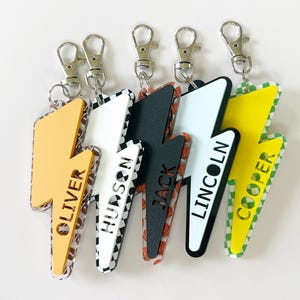 May include: A collection of personalized lightning bolt keychains with names like Oliver, Hudson, Jack, Lincoln, and Cooper. Each keychain features a different design, including sports ball patterns and solid colors, with silver-colored clasps.