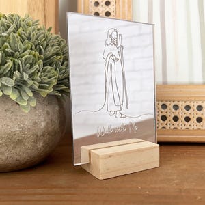 May include: A clear acrylic sign with a line drawing of a figure holding a staff, set in a wooden stand. The words "Walk with Me" are etched below the figure. A faux plant in a stone pot is to the left.