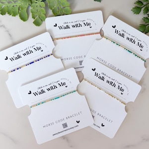 May include: A collection of Morse code bracelets displayed on white cards. Each bracelet features a different color scheme, with beads arranged to spell out a message. The cards read "Walk with Me" and "Morse Code Bracelet".