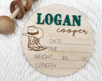 May include: A round wooden baby announcement sign with the name "LOGAN" in green 3D letters and the last name "cooper" in a cursive font. A cowboy hat and boot graphic is etched on the wood. Includes lines for date, time, weight (lbs, oz), and length.