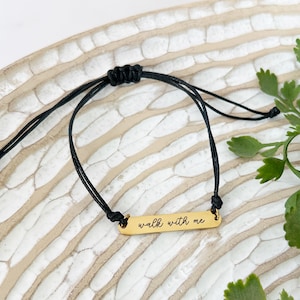 May include: A gold-colored bar bracelet with the phrase "walk with me" in cursive script. The bracelet is strung on a black cord, and the background is a light-colored, textured surface with green foliage.