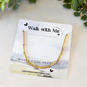 Könnte beinhalten: Goldfarbene Morsecode-Halskette, präsentiert auf einer weißen Karte mit dem Text "Walk with Me" und "Morse Code Necklace". Die Halskette besteht aus zylindrischen und kugelförmigen Perlen. Die Karte enthält auch die Website pennylanecreative.etsy.com.
