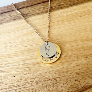 Könnte beinhalten: Eine goldfarbene Halskette mit einer Kette und zwei runden Anhängern. Der obere Anhänger ist silberfarben mit einer Kartenkontur und dem Buchstaben "H". Der untere Anhänger ist goldfarben und trägt die Aufschrift "MISSIONARY MOM".