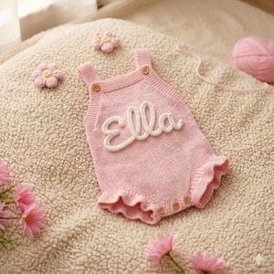 Könnte beinhalten: Ein hellrosa gestrickter Strampler mit gerüschtem Saum und zwei Schulterträgern mit kleinen Holzknöpfen. Der Name Ella ist in weißer Schreibschrift auf die Brust gestickt. Das Kleidungsstück ist mit zwei kleinen gestrickten Blumen verziert.