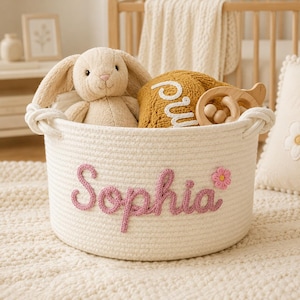 May include: A round white woven rope storage basket featuring the name Sophia embroidered in pink yarn on the front, accented with a small pink flower. Inside the basket sits a beige plush rabbit, a mustard yellow knit item, and a wooden toy.
