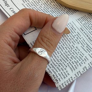 May include: A silver signet ring with an oval face, engraved with the letter "C". The ring is worn on a finger, showcasing its polished finish. The ring is a classic piece of jewelry.