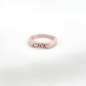 Op de afbeelding: Een ros&eacute;gouden ring met de gegraveerde letters "CHK". De ring heeft een rechthoekige voorkant en een gladde, gepolijste afwerking. De ring wordt gepresenteerd op een wit oppervlak.