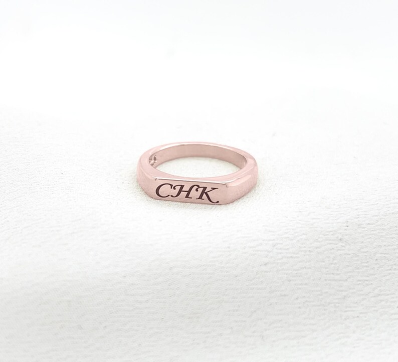 Op de afbeelding: Een ros&eacute;gouden ring met de gegraveerde letters "CHK". De ring heeft een rechthoekige voorkant en een gladde, gepolijste afwerking. De ring wordt gepresenteerd op een wit oppervlak.