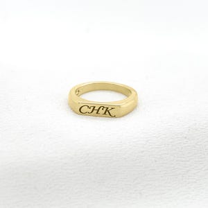 Op de afbeelding: Een goudkleurige ring met de gegraveerde letters "CHK" op een rechthoekig vlak. De gepolijste ring staat tegen een witte achtergrond. Dit eenvoudige sieraad is een tijdloos accessoire, geschikt voor dagelijks gebruik.