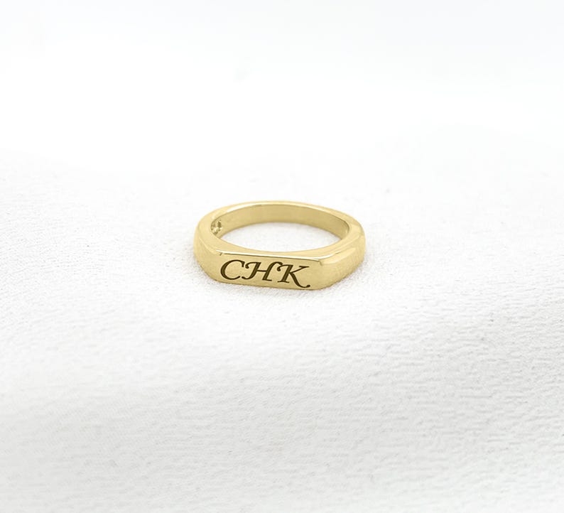 Op de afbeelding: Een goudkleurige ring met de gegraveerde letters "CHK" op een rechthoekig vlak. De gepolijste ring staat tegen een witte achtergrond. Dit eenvoudige sieraad is een tijdloos accessoire, geschikt voor dagelijks gebruik.