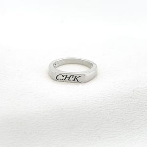 Op de afbeelding: Een zilveren ring met de gegraveerde letters "CHK" op een rechthoekig vlak. De ring heeft een gladde, gepolijste afwerking en een afgeronde band. De ring wordt gepresenteerd op een wit, getextureerd oppervlak.