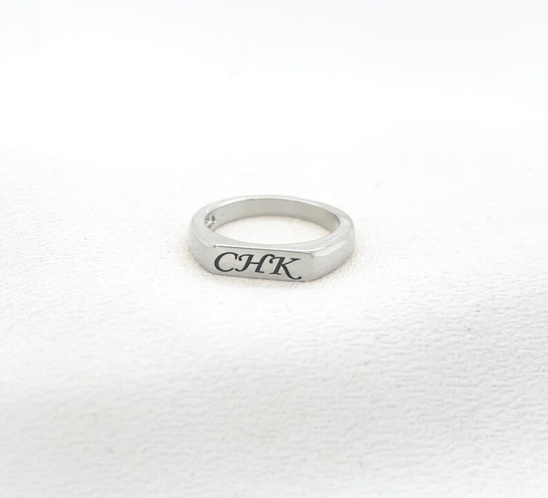 Op de afbeelding: Een zilveren ring met de gegraveerde letters "CHK" op een rechthoekig vlak. De ring heeft een gladde, gepolijste afwerking en een afgeronde band. De ring wordt gepresenteerd op een wit, getextureerd oppervlak.