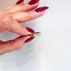 Op de afbeelding: Een gouden ring met de letters "CHK" gegraveerd. De ring wordt vastgehouden tussen vingers met lange, gepolijste, bordeauxrode nagels. De achtergrond is een effen wit oppervlak.