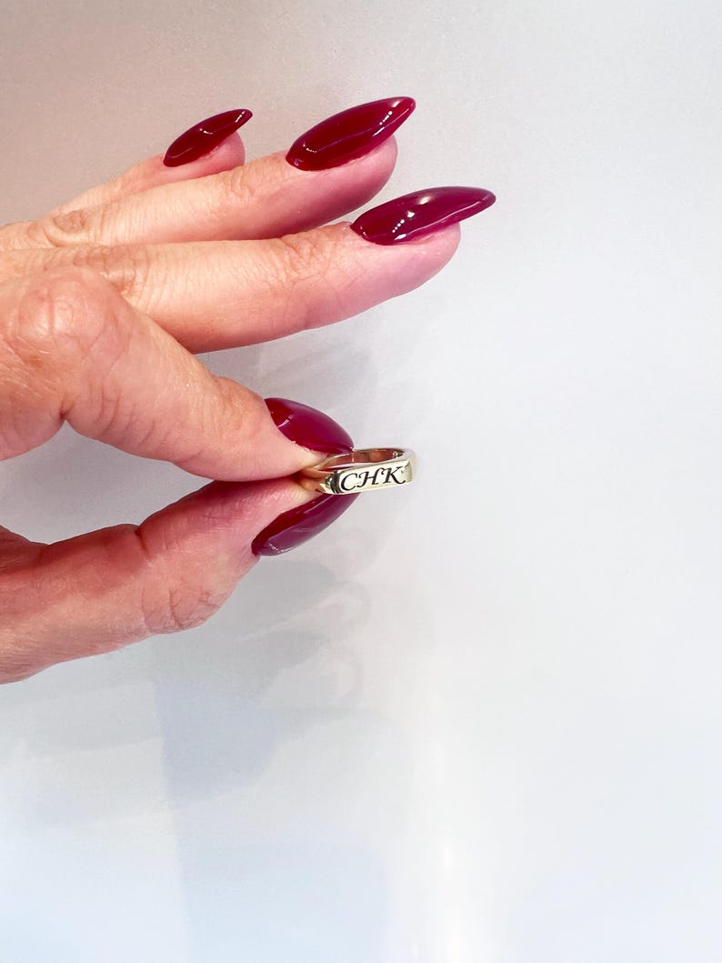 Op de afbeelding: Een gouden ring met de letters "CHK" gegraveerd. De ring wordt vastgehouden tussen vingers met lange, gepolijste, bordeauxrode nagels. De achtergrond is een effen wit oppervlak.
