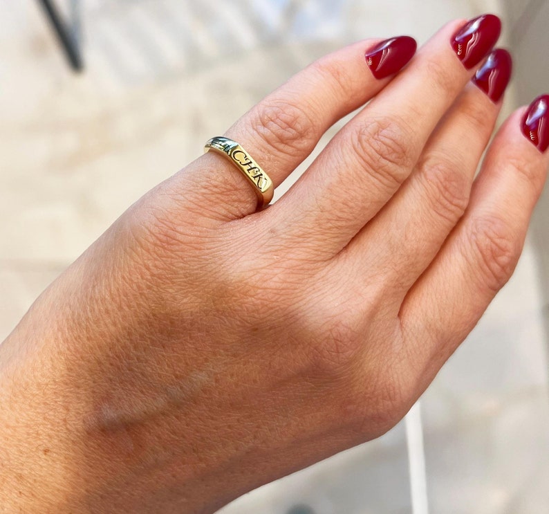 Op de afbeelding: Een gouden ring met de letters "CHK" gegraveerd op de band. De ring wordt om een vinger gedragen en de hand heeft rode nagellak. De ring lijkt van goud te zijn en heeft een gladde, gepolijste afwerking.