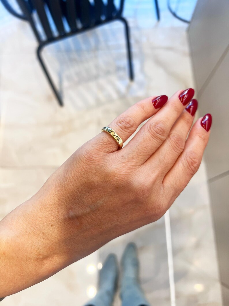 Op de afbeelding: Een gouden ring met het woord "CHER" gegraveerd. De ring zit aan een vinger met rode nagellak. De achtergrond is een lichtgekleurde tegelvloer en een zwarte stoel.