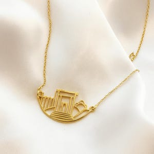 May include: A gold-colored necklace featuring a stylized architectural design. The pendant depicts a building with columns and a curved base, suspended from a delicate chain. The necklace is displayed on a white, textured surface.