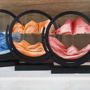 May include: Three circular sand art displays with black frames and rectangular bases. Each piece contains coloured sand forming mountain-like landscapes in blue, orange, and red, layered between clear glass panels with visible air bubbles.