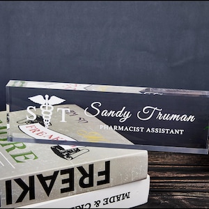 Puede incluir: Una placa de escritorio de acrílico transparente con el nombre "Sandy Truman" y el título "Asistente de Farmacia" en letras blancas. La placa también presenta un símbolo médico y las iniciales "S T".