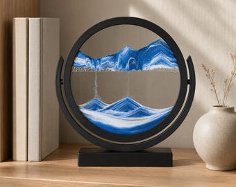 May include: A circular black frame holds a glass display filled with layered blue, white, and gold sand. The sand forms mountain-like peaks and valleys within the rotating frame, which is mounted on a rectangular black base for stability.