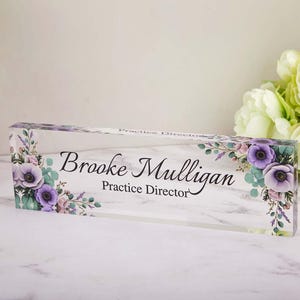 May include: A clear acrylic desk nameplate featuring the name "Brooke Mulligan" and the title "Practice Director" in black script. The nameplate is decorated with purple and pink floral accents.