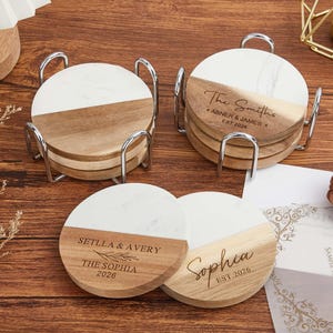 Puede incluir: Un juego de posavasos redondos con un diseño dividido de mármol blanco y madera natural. Dos pilas se mantienen en soportes de metal plateado. Las secciones de madera están grabadas con nombres y fechas, incluyendo The Smiths, Abner & James, Setlla & Avery y Sophia.