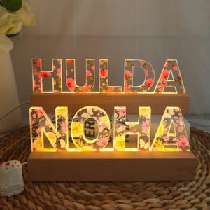 Op de afbeelding: Twee verlichte houten naamborden. Het bovenste bord spelt "HULDA" en het onderste bord spelt "NOHA". De letters zijn helder met een bloemenontwerp en worden van binnenuit verlicht. De borden staan op houten voetstukken.