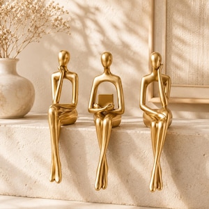 Peut inclure: Trois figurines humaines abstraites de couleur dorée en position assise. La figurine de gauche pose une main sur son menton, celle du centre tient un objet rectangulaire et celle de droite pose une main sur sa joue. Surfaces lisses.