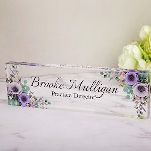 May include: A clear acrylic desk nameplate featuring the name "Brooke Mulligan" and the title "Practice Director" in black script. The nameplate is decorated with purple and pink floral accents.
