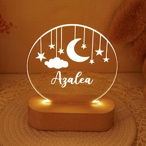 May include: A glowing night light with a wooden base. The clear acrylic design features a crescent moon, stars, and the name "Azalea" in script. The light emits a warm, inviting glow, perfect for a nursery or bedroom.
