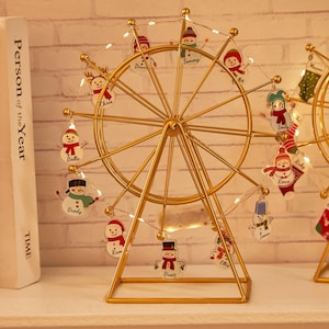 Peut inclure: Une décoration de Noël en forme de grande roue en métal doré. La roue présente de petits ornements de bonhommes de neige avec des noms et est illuminée par de petites lumières. La base est de forme géométrique simple.