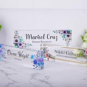 Op de afbeelding: Drie rechthoekige bureau-naamplaatjes van helder acryl met bloemmotieven in tinten paars, blauw en roze. De plaatjes tonen de namen Marisol Cruz, Cherre Wright en Nikki Gilbert, samen met hun respectievelijke functietitels in zwarte tekst.