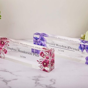 Puede incluir: Dos placas de escritorio de acrílico transparente con diseños florales. Una presenta flores rosas y burdeos, la otra moradas. Cada una muestra un nombre y credenciales en texto negro: "Daphne Henderson PhD LCSW" y "Shauna Hayden-Zbranek LCSW".