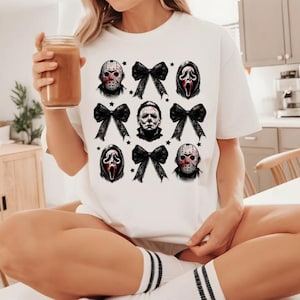 May include: White t-shirt featuring a design with horror movie characters and black bows. The design includes images of Jason Voorhees, Michael Myers, and Ghostface, along with black bows and stars. The shirt is a casual, crew neck style.