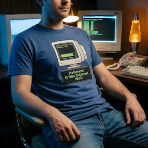 May include: A blue short-sleeved t-shirt featuring a graphic of a vintage computer monitor with a loading bar at 95 percent. Below the monitor, a black rectangular box contains the green text: Patience: A 90s Internet Skill.