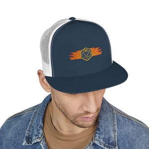 May include: A navy blue trucker hat with a white mesh back and a flat peak. The hat features an embroidered gold and orange design of a 20-sided die with a burst of flames. The hat is worn by a person wearing a denim jacket.