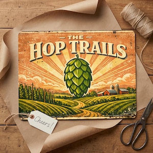 Può includere: Un cartello in metallo in stile vintage con la scritta "THE HOP TRAILS" e un'illustrazione di un grande cono di luppolo verde. Il cartello presenta un paesaggio rurale con campi, una strada e un fienile rosso. Un'etichetta con "Cheers!" è allegata.