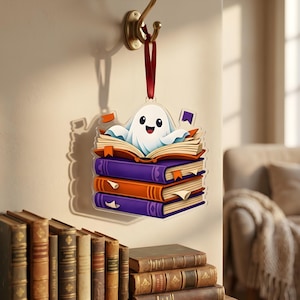 May include: A Halloween decoration showing a cartoon ghost character perched on a pile of books. The books are coloured purple and orange, and the ornament hangs from a red ribbon. The ghost has a happy face and is surrounded by open books.