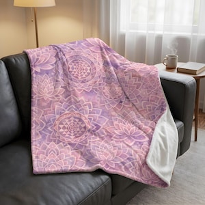 May include: A soft, plush blanket in shades of lavender and pink, featuring a repeating pattern of lotus flowers and geometric designs. The blanket is draped over a black leather couch, with a white, fluffy underside visible.