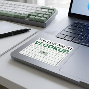 May include: A rectangular sticker featuring the text You Had Me At VLOOKUP in dark green letters above a grid graphic with columns labelled A through E and a small black heart icon in one of the cells, applied to the surface of a laptop computer.