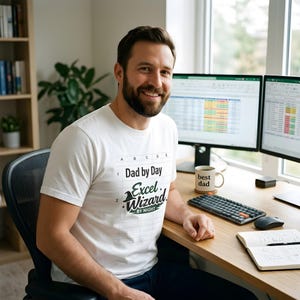 May include: A man wearing a white t-shirt with the text "Dad by Day Excel Wizard by Night." He is sitting at a desk with two computer monitors, a coffee mug that says "best dad," a keyboard, and a notebook.