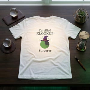 May include: A white short-sleeve t-shirt featuring the text Certified XLOOKUP Sorcerer in dark gray. Below the text is a graphic of a green pie chart wearing a purple witch hat with a yellow band, surrounded by small decorative star icons.